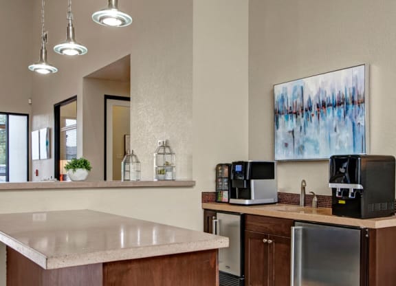 Modern office breakroom here at Bolero Apartments with a coffee station, soft lighting, abstract wall art, and a stone countertop. The ambiance is clean and inviting.