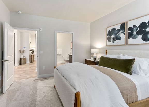 A serene bedroom here at Reflect at Dobson Ranch Apartments with a plush bed adorned with white and beige linens and a green pillow. Minimalist decor includes floral artwork and a wooden nightstand with a lamp. An open door leads to a hallway with light wooden floors, decorated with vases and a mirror.
