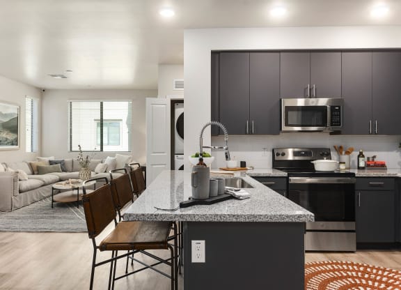 Modern open-plan kitchen and living room here at Reflect at Dobson Ranch Apartments. Gray kitchen cabinets and granite island with brown stools. Cozy cream sofa in lounge area with large windows.