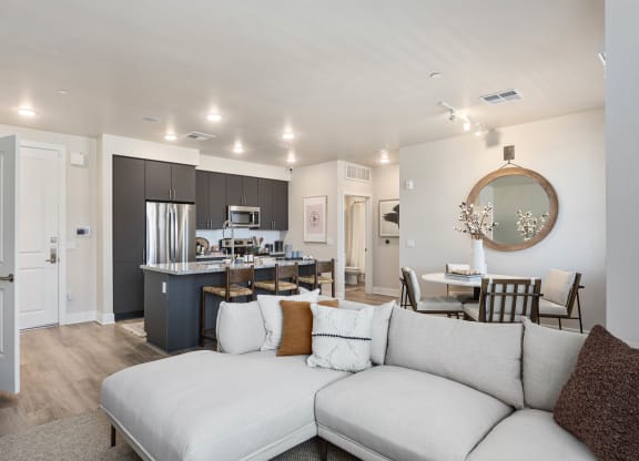 Modern open-plan living area here at Reflect at Dobson Ranch Apartments with a cozy beige sectional sofa, a sleek kitchen with dark cabinets, a round dining table, and warm lighting.
