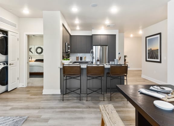 Modern kitchen here at Reflect at Dobson Ranch Apartments with sleek dark cabinets, stainless steel appliances, and a central island with three brown stools. Dining table set nearby. Warm and inviting.