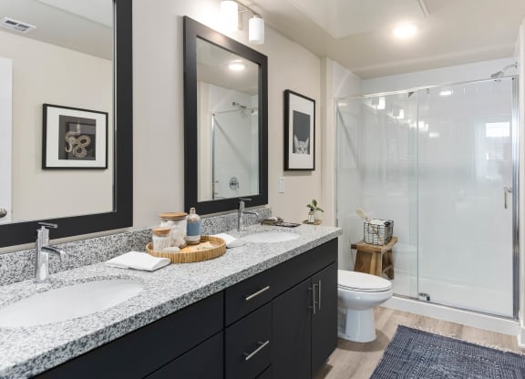 Modern bathroom here at Reflect at Dobson Ranch Apartments with two sinks on a granite countertop, large mirrors, and a glass-enclosed shower. Decor includes framed art and subtle plants.