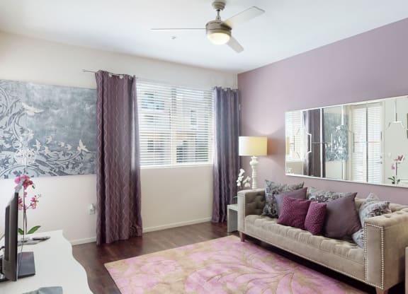 Modern living room here at Artisan at Main Street Metro Apartments with lavender walls, grey tufted sofa, and pink cushions. Large mirror, abstract art, and floral rug complement the chic decor.