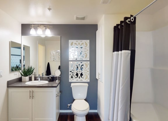 A modern bathroom here at Artisan at Main Street Metro Apartments with a sleek design features a white vanity, round basin, and rectangular mirror. There's a toilet, decorative wall art, and a shower with a black and white curtain. The space is bright and organized.