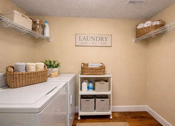 A spacious laundry room here at 3600 Brentwood with a full-size washer and dryer, open wire shelving for storage, and warm wood-style flooring for a clean, convenient setup.