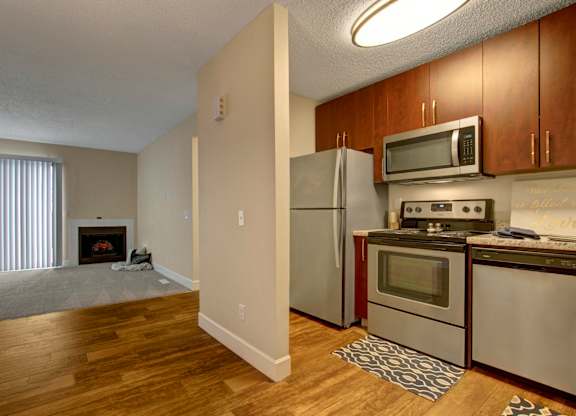 A spacious open-concept living area here at 3600 Brentwood with warm wood-style flooring, an updated kitchen with stainless steel appliances, and a cozy living room featuring a fireplace and tall sliding doors with vertical blinds.
