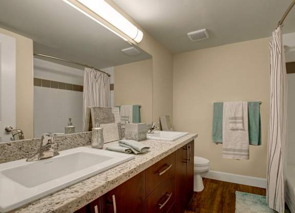 A modern bathroom here at 3600 Brentwood with a double-sink vanity, granite-style countertops, and a large mirror, featuring a bathtub-shower combo and clean, bright lighting.