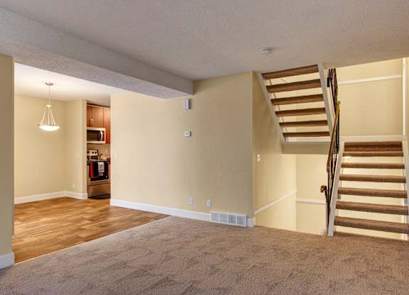 A spacious, open-concept living area here at 3600 Brentwood with soft carpet, warm wood-style flooring in the dining space, and a staircase leading to the upper level, with the kitchen visible just beyond.