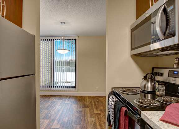 A modern kitchen here at 3600 Brentwood with stainless steel appliances, an electric stove, and granite-style countertops, opening into a bright dining area with wood-style flooring and tall windows with vertical blinds.