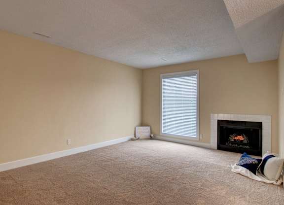 A spacious, empty living room here at 3600 Brentwood with soft beige carpet, a bright window with white blinds, and a cozy fireplace that adds warmth to the open space.