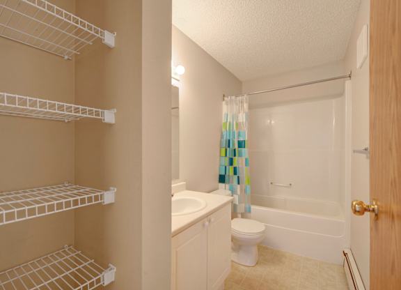 A clean bathroom here at Aspen Terrace with a bathtub-shower combo, a white vanity with a smooth countertop, and a bright mirror light. A built-in shelving nook with wire racks provides extra storage, giving the space a practical, fresh feel.