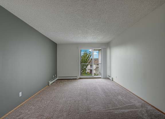 A spacious, empty living room here at Aspen Terrace with soft carpet, neutral walls, and a sliding glass door that opens to a private balcony. A large window and bright natural light create an airy, inviting feel.