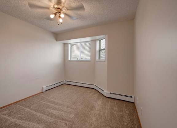 A bright, empty bedroom here at Aspen Terrace with beige carpet, neutral walls, and a ceiling fan with overhead lighting. A corner window with blinds brings in natural light, creating a cozy, comfortable feel.
