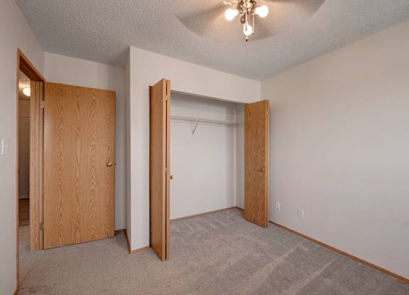 A bright, empty bedroom here at Aspen Terrace with soft carpet, neutral walls, and a ceiling fan with overhead lighting. A roomy closet with wood doors provides convenient storage, creating a clean, comfortable feel.