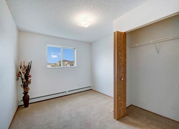 A bright bedroom here at Aspen Terrace with beige carpet, neutral walls, and a window that brings in natural light. A spacious open closet with wood doors and a hanging shelf adds convenient storage, creating a clean, comfortable feel.