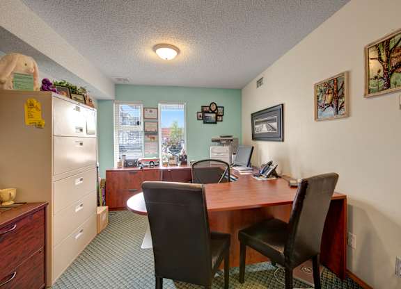 A welcoming office area here at Aspen Terrace with a spacious wood desk, guest chairs, and natural light from front windows. A tidy workspace with filing cabinets, computer setup, and warm wall décor creates a professional, friendly feel.