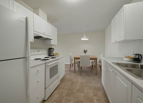 A spacious kitchen and dining area here at Aspen Terrace with white cabinets, matching white appliances, and a double stainless steel sink on wide countertops. A simple dining table with upholstered chairs sits under a modern pendant light, giving the room a clean, bright feel.