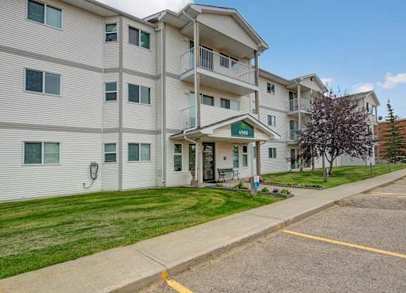 A welcoming exterior entrance here at Aspen Terrace featuring a well-maintained apartment building with private balconies and landscaped green lawns. A covered entryway with building signage and nearby parking creates a convenient, friendly community feel.