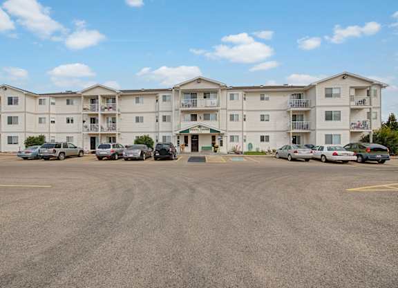 A wide exterior view here at Aspen Terrace showcasing a large, well-maintained apartment building with private balconies and a rental center entrance. A spacious parking area sits out front under a bright blue sky, creating an open, welcoming community feel.