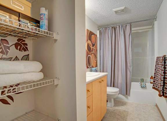 A functional bathroom here at Aspen Terrace with a bathtub-shower combo, a light wood vanity with a white countertop, and a clean tile floor. A built-in shelving area with wire racks adds convenient storage, giving the space a practical, welcoming feel.