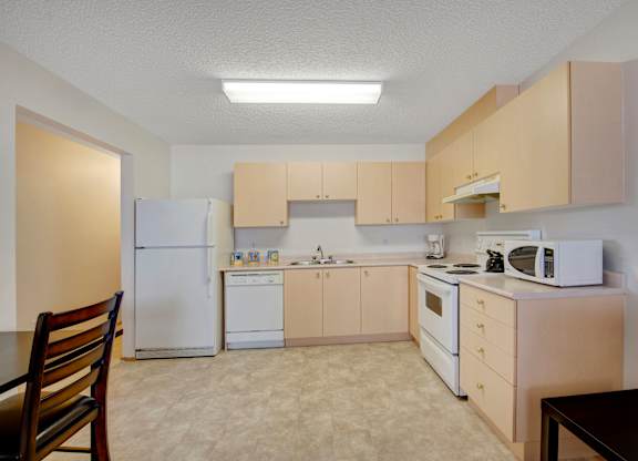 A spacious kitchen here at Aspen Terrace with light wood cabinets, white countertops, and matching white appliances including a refrigerator, stove, and dishwasher. A double sink and open floor space create a clean, bright layout with a simple, neutral feel.