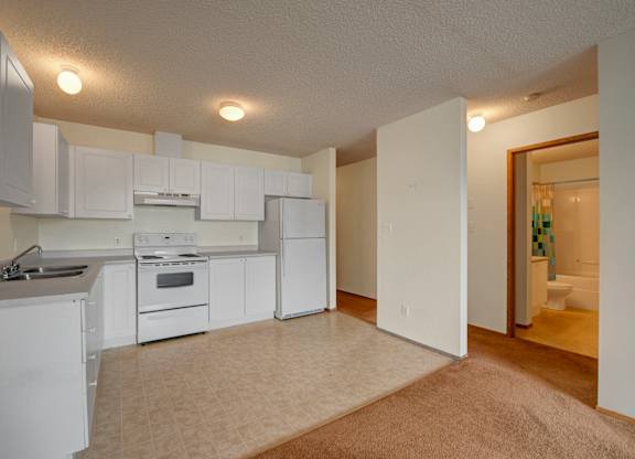 A bright open kitchen here at Aspen Terrace with white cabinets, white appliances, and a double stainless steel sink on spacious countertops. A wide layout with tiled flooring and an adjoining carpeted living area creates an open, comfortable feel.