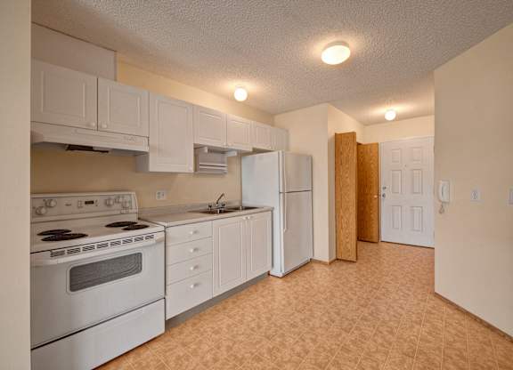 A bright kitchen here at Aspen Terrace with white cabinets, white appliances, and a stainless steel sink with generous counter space. A wide tiled floor and overhead lighting create an open, practical layout with a clean, neutral feel.