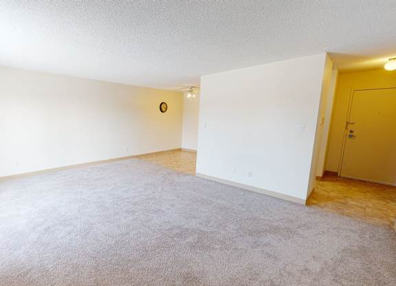 A bright open living room here at Cambridge West with soft carpet flooring, crisp white walls, and a wide layout with plenty of open space. Large windows with vertical blinds bring in natural light, while the entry hallway with tile flooring and warm overhead lighting adds a practical flow into the home.