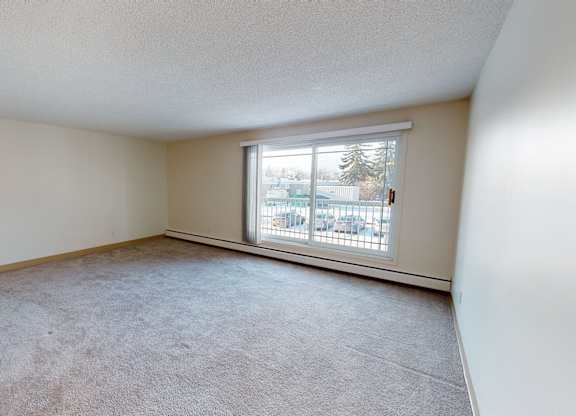 A spacious, empty living room here at Cambridge West with soft beige carpet and light neutral walls, brightened by natural light from a large sliding glass door that opens to a balcony with outdoor views.