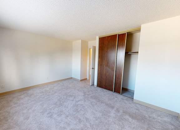 A bright, empty bedroom here at Cambridge West with soft beige carpet and light neutral walls, featuring a large window for natural light and a spacious closet with sliding wood doors for convenient storage.