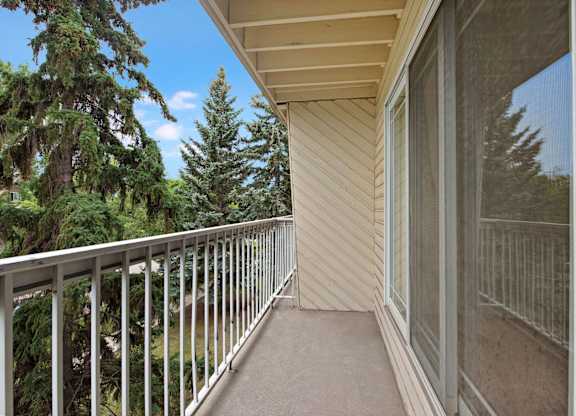 A private balcony here at Cambridge West with a covered outdoor space, sturdy railing, and peaceful views of mature evergreen trees, accessed through a large sliding glass door.