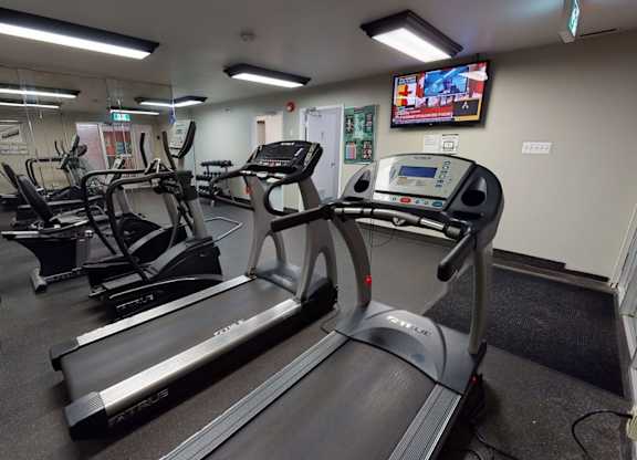 A well-equipped fitness room here at Cambridge West with treadmills, cardio machines, and free weights, featuring mirrored walls, rubber flooring, and bright lighting with a mounted TV for added convenience.