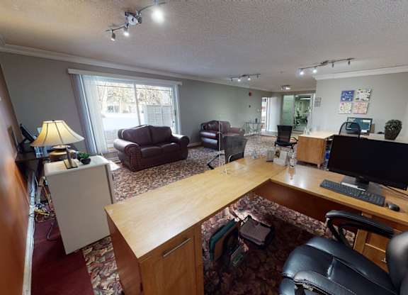 A cozy community lounge here at Cambridge West with comfortable leather seating, patterned carpet, and a bright business center setup with desks and computers, plus sliding glass doors that bring in natural light and outdoor views.