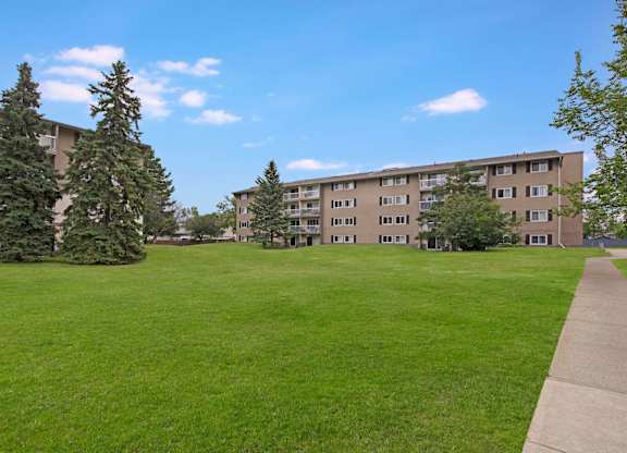 A wide outdoor green space here at Cambridge West with open grassy areas, mature trees, and a paved walkway, offering a peaceful setting in front of the apartment buildings with balcony views.