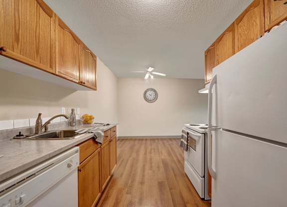 A bright kitchen and dining space here at Cambridge West with warm wood cabinets, white appliances, and a long countertop with a stainless sink and dishwasher. Wood-style flooring, a ceiling fan, and an open dining area with a wall clock create a simple, functional layout with plenty of room to move around.