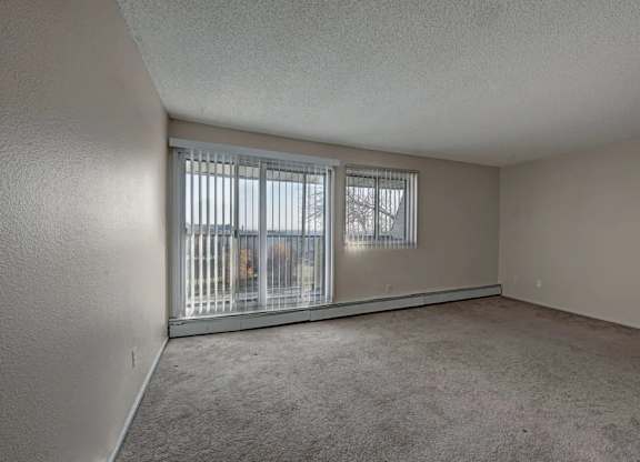 A bright living room here at Cedar Gardens with a wide open layout and soft carpeting, featuring large sliding glass doors with vertical blinds, an extra window for added natural light, and a long baseboard heater along the wall for a cozy, comfortable feel.
