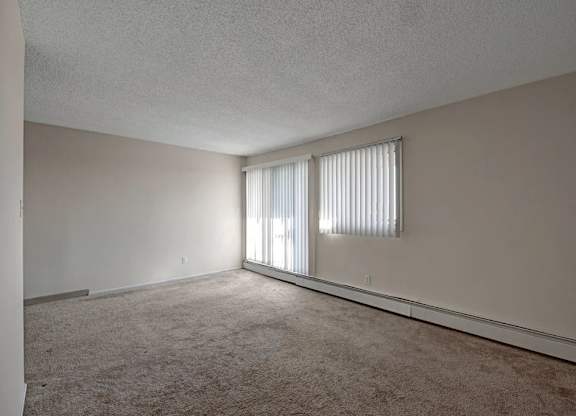 An airy living room here at Cedar Gardens with plush carpeting and a long open layout, featuring a sliding glass door with vertical blinds, an additional window for natural light, and a baseboard heater along the wall for a warm, comfortable feel.