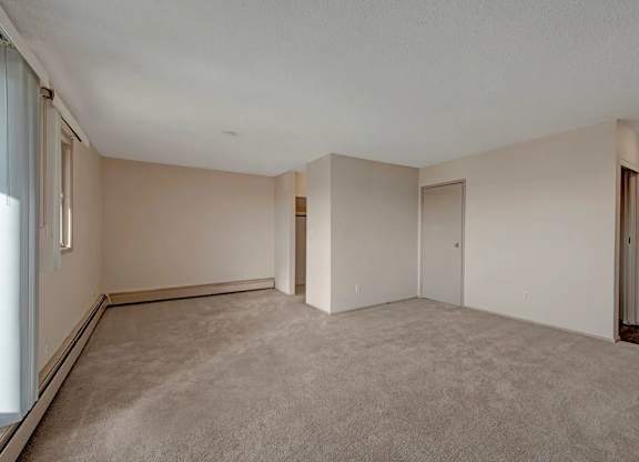 A spacious living area here at Cedar Gardens with soft carpeting and a bright, open layout, featuring a large window with vertical blinds, a long baseboard heater along the wall, and a versatile nook near the entryway for a cozy, comfortable feel.