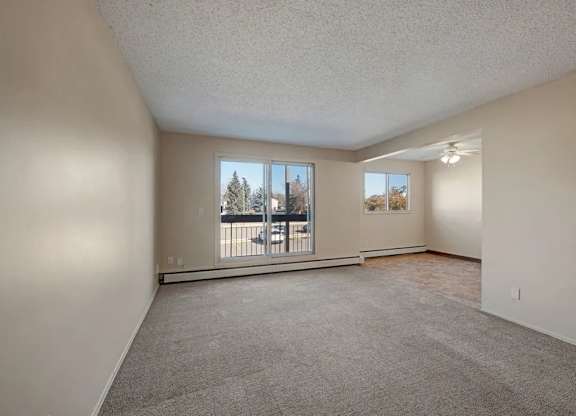 A bright open living room here at Cedar Gardens with soft carpeting and a wide layout, featuring a large sliding glass door for natural light, a baseboard heater along the wall, and a connected dining nook with an additional window and ceiling fan for an airy, comfortable feel.