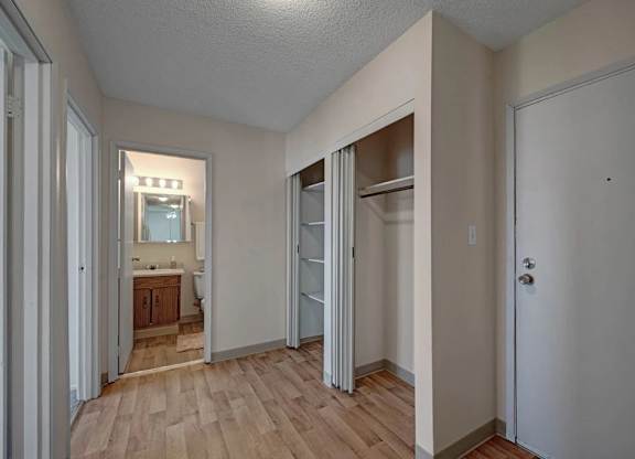 A welcoming entryway here at Cedar Gardens with wood-style flooring and a clean, neutral look, featuring a convenient coat closet with shelving, a wide hallway for easy access, and a nearby bathroom vanity with bright lighting for an everyday-ready setup.
