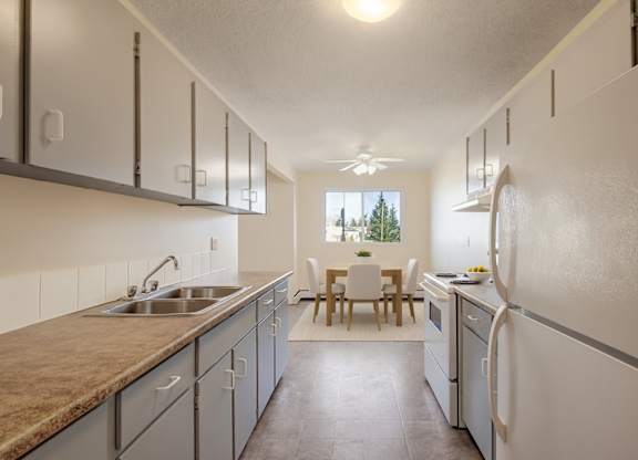 A bright kitchen here at Cedar Gardens with plenty of cabinet storage and generous counter space, featuring a double sink, white appliances, and an open dining area with a cozy table set by the window for a clean, comfortable feel.