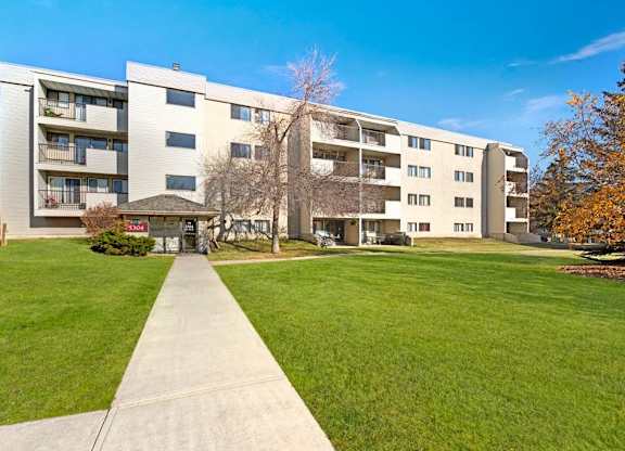 A welcoming exterior view here at Cedar Gardens with a well-kept multi-level apartment building, featuring private balconies, a wide concrete walkway to the entrance, and a large green lawn with mature trees for a bright, open feel.