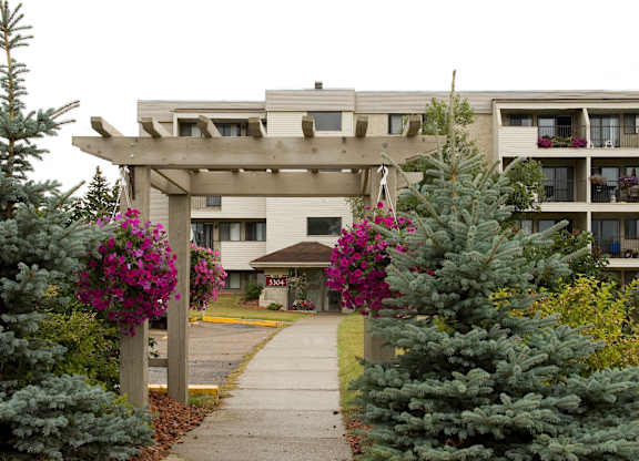 Here at Cedar Gardens, a charming pergola entryway with bright purple hanging flower baskets leads to the apartment building, surrounded by lush evergreen trees, green shrubs, and a clean walkway up to the front entrance.
