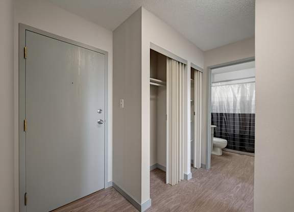 A bright entryway here at Cedar Gardens with wood-style flooring and a sleek front door, featuring convenient closet storage with sliding doors, clean neutral walls, and a simple hallway layout that leads easily into the bathroom for an open, welcoming feel.