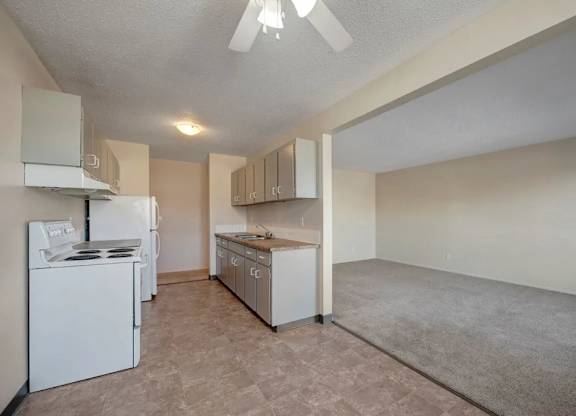 A bright open-concept space here at Cedar Gardens with a functional galley kitchen and adjoining living area, featuring white cabinetry, full-size appliances, and durable flooring that flows into soft carpeting for a cozy, everyday feel.