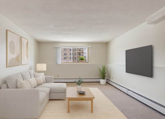 A bright living room here at Gateway Square featuring a cozy light-gray sectional with soft neutral pillows, paired with modern wall art, a warm wood coffee table on a plush area rug, and a few simple plants for a clean, relaxing feel.
