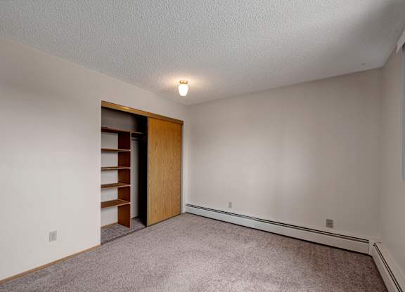A spacious, empty bedroom here at Gateway Square with soft beige walls, plush carpet flooring, and a large closet featuring sliding wood doors and built-in shelving for convenient storage.
