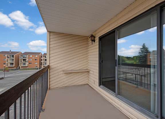 A private outdoor balcony here at Gateway Square with durable flooring, a sliding glass door leading inside, and open views of nearby buildings and greenery under a bright sky.