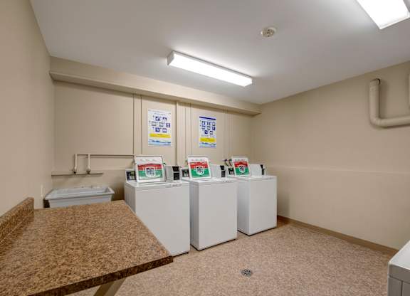 A clean, well-lit laundry room here at Gateway Square with multiple top-loading washers, neutral walls, durable flooring, and a spacious folding counter for added convenience.
