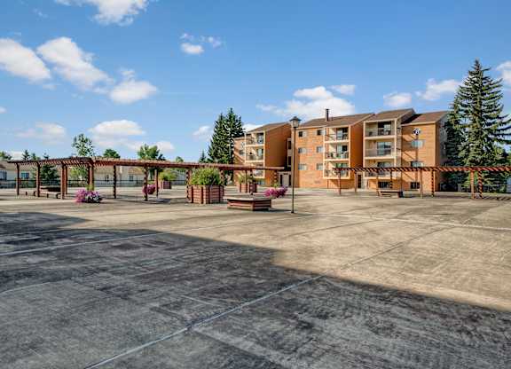 A spacious outdoor courtyard here at Gateway Square with open concrete walkways, wooden pergolas, landscaped planters, and surrounding apartment buildings under a bright blue sky.